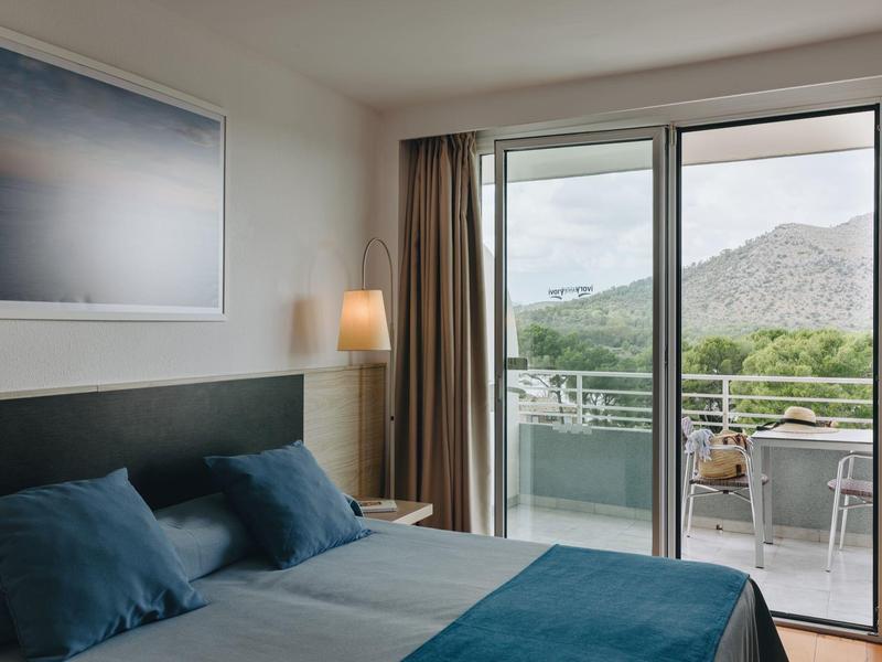 Dormitorio con cama doble, cojines y manta azules, acceso a balcón con vista a la montaña.