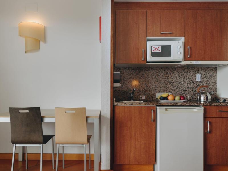 Pequeña cocina de hotel con microondas, nevera y mesa con dos sillas de diferentes colores.