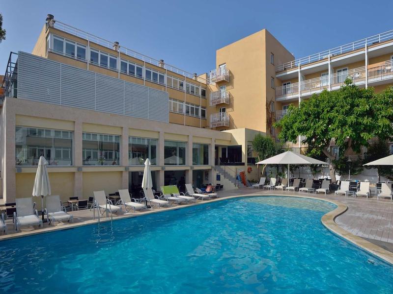 Hotel z dużym basenem na zewnątrz, leżakami i parasolami pod czystym niebieskim niebem.