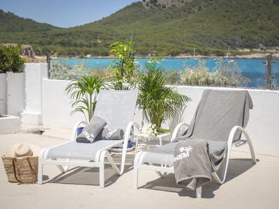 Zwei weiße Sonnenliegen mit grauen Handtüchern auf einer sonnigen Terrasse mit Blick auf das Meer und grüne Hügel.