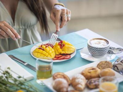 Person schneidet Mangoscheiben auf einem Teller mit verschiedenen Früchten neben Gebäck und Getränken.