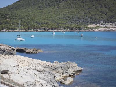 Klares blaues Wasser mit Booten an einer felsigen Küste und bewaldetem Hügel im Hintergrund