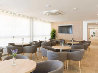 Salone luminoso con tavoli rotondi e poltrone grigie, decorazione semplice con pianta verde.