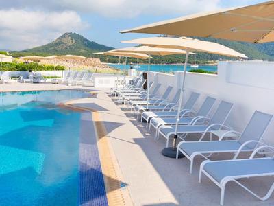 Lettini bianchi allineati accanto a una piscina con ombrelloni, montagna sullo sfondo.