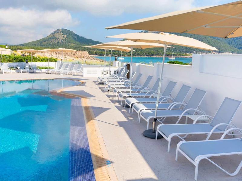 Lettini bianchi allineati accanto a una piscina con ombrelloni, montagna sullo sfondo.