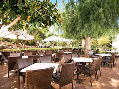 Terrassenbereich mit braunen Stühlen, weißen Tischtüchern und Sonnenschirmen unter Bäumen.