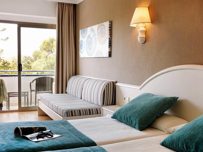 Jasny pokój hotelowy z dwoma łóżkami, kanapą, balkonem i widokiem na drzewa w świetle dziennym.