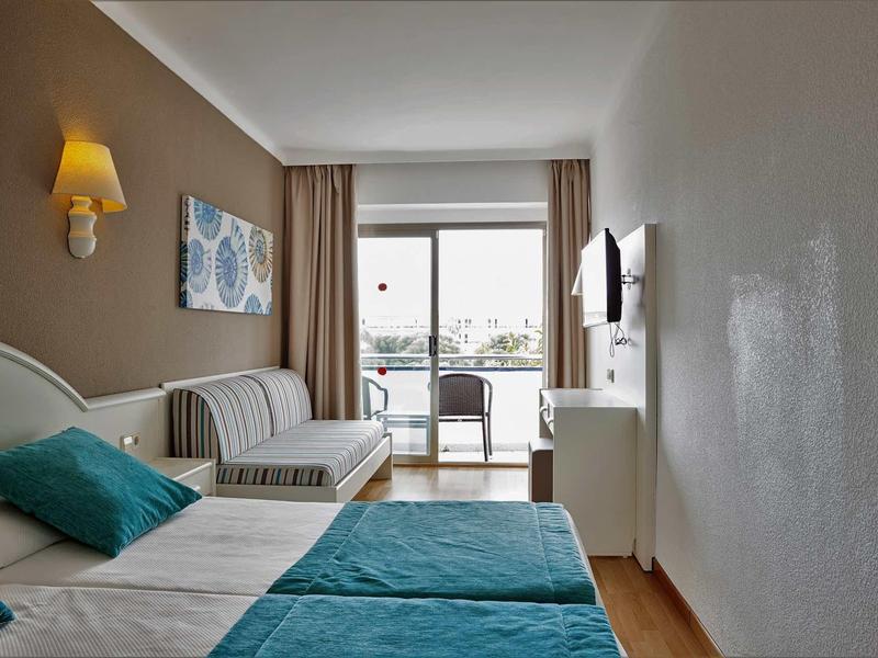 Jasny pokój hotelowy z podwójnym łóżkiem, sofą, biurkiem i balkonem z widokiem na zewnątrz.