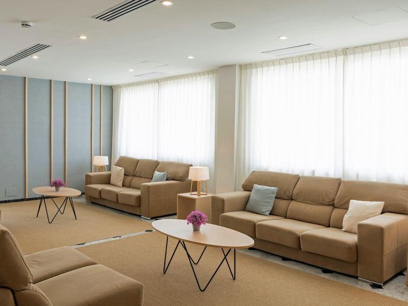 Bright hotel lounge with beige sofas, coffee tables, lamps, and large windows with sheer curtains.