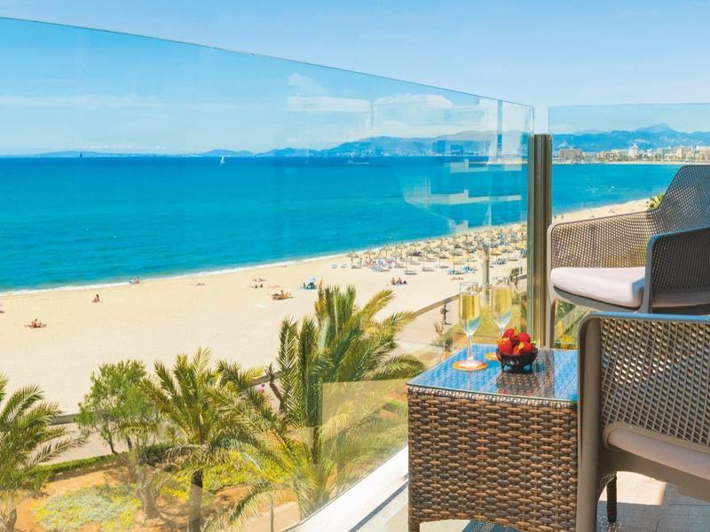 Balcony with chairs overlooking a sunny beach, palm trees, and a calm blue sea.