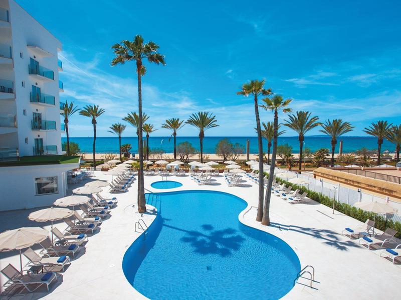 Outdoor pool area with lounge chairs, umbrellas, palm trees, and ocean view at a hotel.