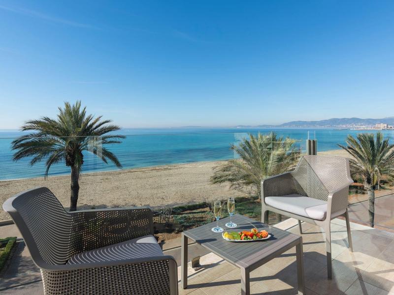 Ocean view from a terrace with two chairs, a table with food, palm trees, and a clear blue sky.