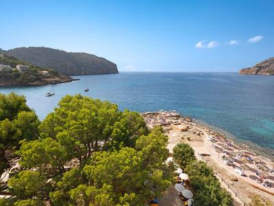 Klares Meer, Sandstrand mit Liegen, grüne Kiefern, Hügel im Hintergrund, blauer Himmel.