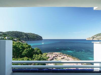 Blick von Balkon mit weißem Geländer auf Meer, Sandstrand und bewaldete Hügel unter blauem Himmel.