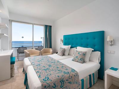 Modernes Hotelzimmer mit Doppelbett, türkisfarbenem Kopfteil und Meerblick durch große Fenster.