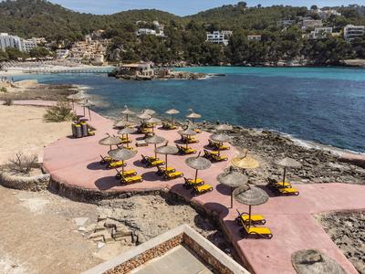 Strand mit gelben Liegen und Strohdach-Sonnenschirmen an einer geschützten Bucht mit klarem Wasser.