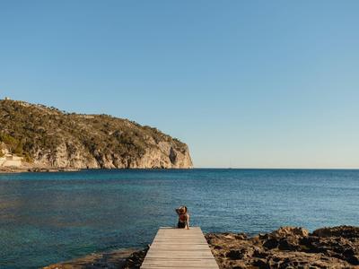 Holzsteg führt ins ruhige blaue Meer, rechts felsige Küste, links bewaldeter Hügel unter klarem Himmel.