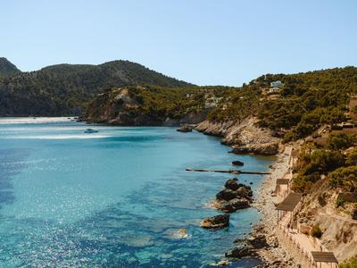 Klares türkisfarbenes Meer trifft auf felsige Küste mit grünen Bäumen und bewachsenen Hügeln im Hintergrund.