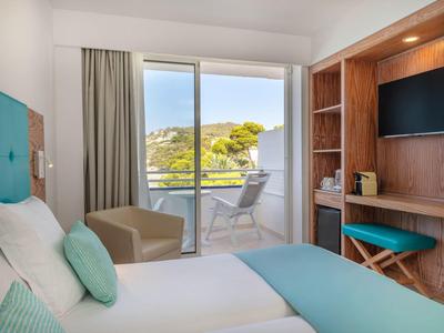 Helles Hotelzimmer mit Bett, Balkon und Blick auf grüne Hügel.