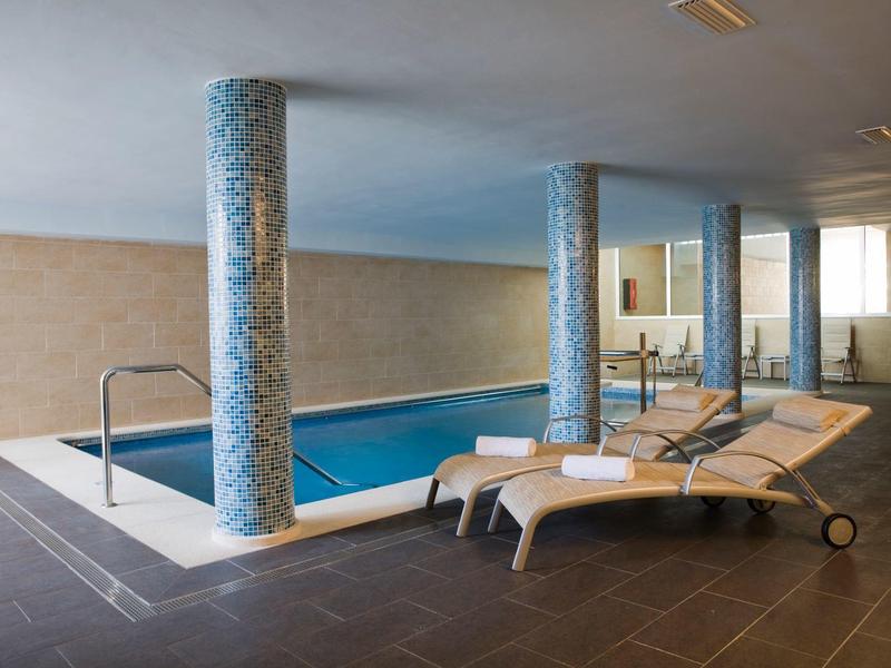 Innenpool mit Mosaiksäulen, Liegestühlen und großen Fenstern, helle Wand und dunkler Boden.