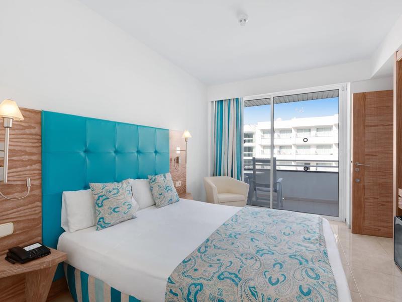 Hell eingerichtetes Hotelzimmer mit großem Bett, blauen Akzenten und Balkon mit Stadtblick.