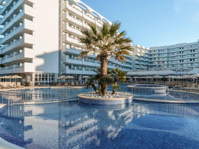 Moderner Hotelkomplex mit großem, geschwungenem Pool und Palmen in der Mitte bei Tageslicht.