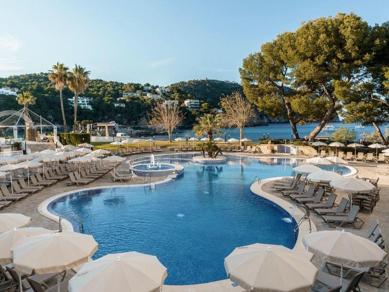 Freizeitpool mit runden Formen, umgeben von Liegestühlen und Sonnenschirmen, vor grünem Hügel und See.