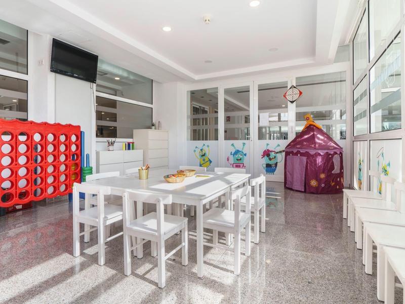 Heller Raum mit weißem Kindertisch, Stühlen, Spielzeug und großem Connect-4-Spiel an der linken Wand.