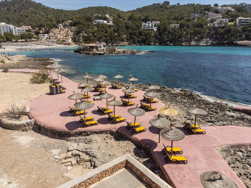 Strand mit gelben Liegen und Strohdach-Sonnenschirmen an einer geschützten Bucht mit klarem Wasser.