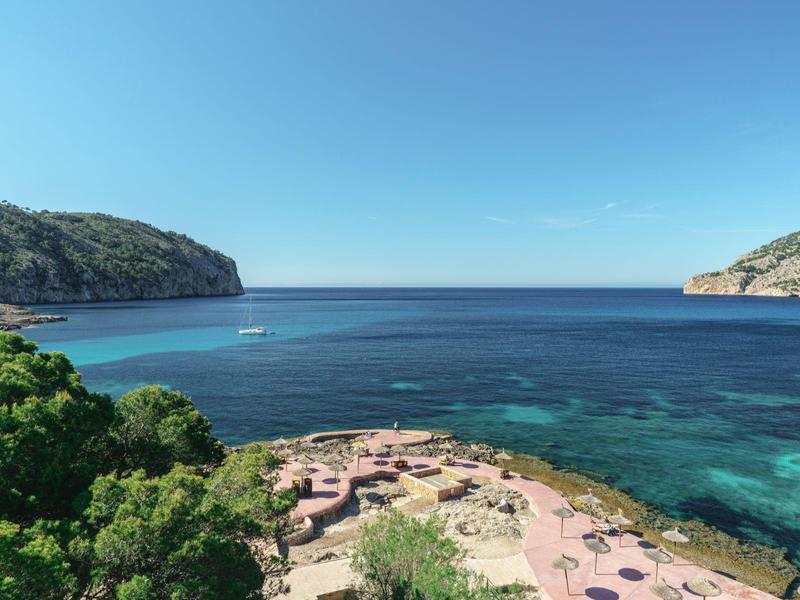 Klares Meer mit felsiger Küste, grün bewachsene Hügel und Sonnenschirme auf Sandstrand.
