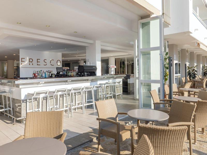 Heller, moderner Hotelbarbereich mit Theke, weißen Barhockern und vielen Sitzgelegenheiten im Innen- und Außenbereich.