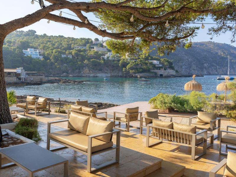 Außenbereich einer Terrasse mit Sitzgelegenheiten und Blick auf eine Bucht und bewaldete Hügel.