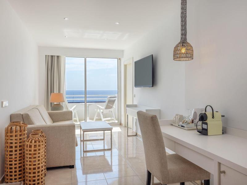 Helles Wohnzimmer mit Sofa, Esstisch, Fernseher und Meerblick durch große Fenster.