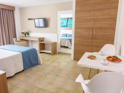 Bright hotel room with double bed, desk, wall-mounted TV, and dining table with snacks.