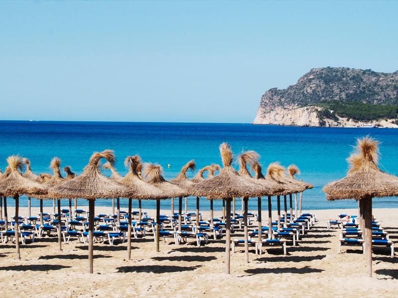 Beach with empty sun loungers under straw roofs and clear blue sea in the background.