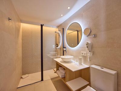Modernes Badezimmer mit Rundspiegel, weißem Waschbecken, Holzregal, Dusche und Toilette in hellem Design.