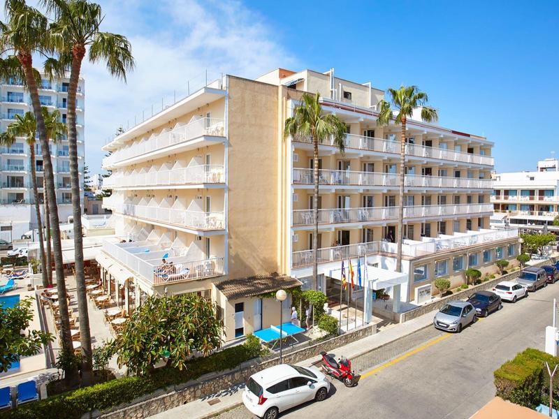 Edificio moderno di un hotel con balconi in una città costiera soleggiata, palme e auto parcheggiate visibili.