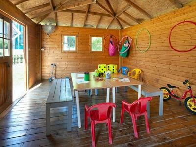 Kinderraum mit Holzwänden, Tischen, bunten Stühlen, Spielzeug und einem Dreirad, hell erleuchtet.
