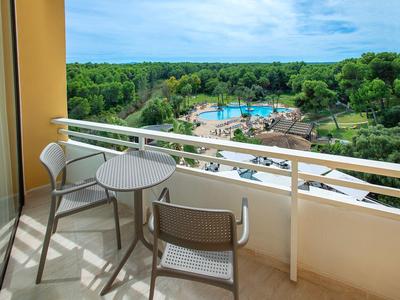 Balkon mit kleinem runden Tisch, zwei Stühlen und Blick auf Pool, grüne Bäume und blauen Himmel.