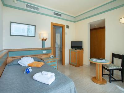 Hotelzimmer mit Einzelbett, Handtüchern, Tisch, Stuhl, Schrank und Lampen in hellem Raum.