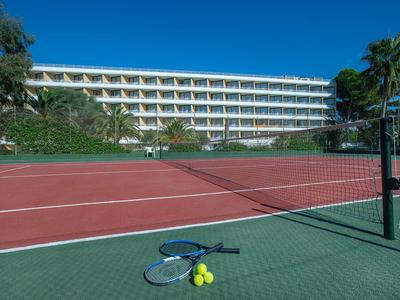 Roter Tennisplatz mit Netz, Tennisschläger und Bällen, im Hintergrund ein mehrstöckiges Hotel.