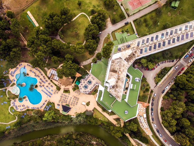 Luftaufnahme eines Resortbereichs mit blauem Pool, Liegestühlen, grünen Wiesen und angrenzender Straße.