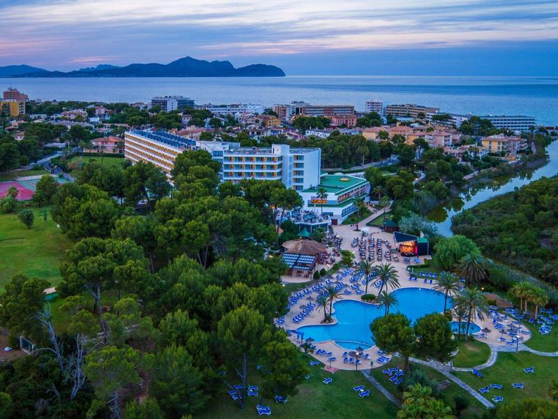 Landschaft mit Hotel, Schwimmbad, viel Grün und Meer im Hintergrund bei blauem Himmel.
