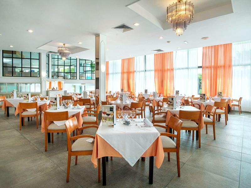 Großes Restaurant mit vielen Tischen, weißen Tischdecken und orangen Vorhängen am Fenster.