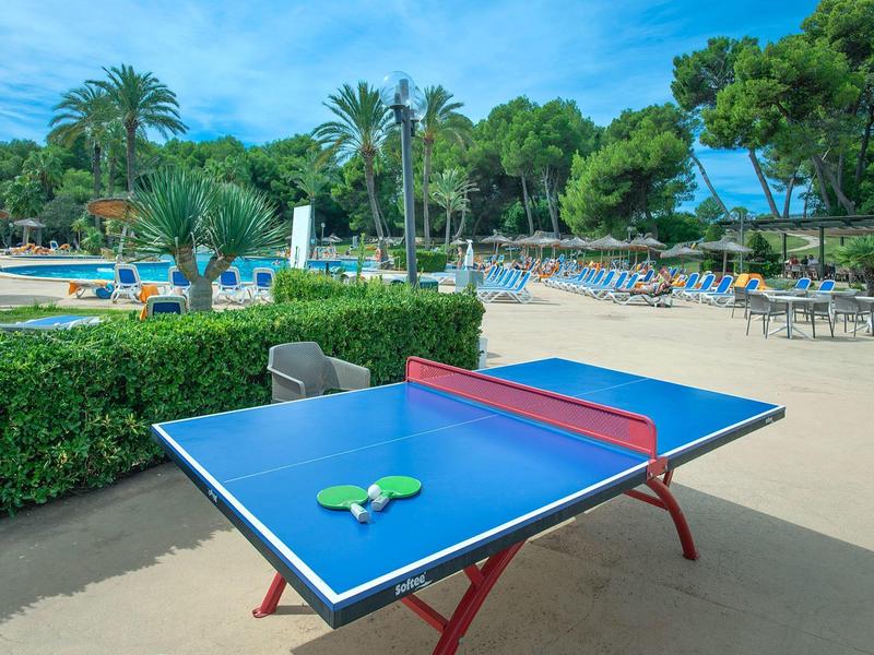 Freizeitbereich mit blauem Tischtennistisch, grünem Busch, Palmen und Sonnenschirmen bei sonnigem Himmel.