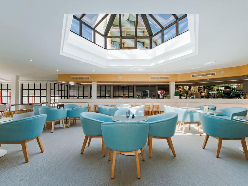 Hell eingerichteter Raum mit blauen Sesseln, Holztischen und großem Oberlichtfenster.