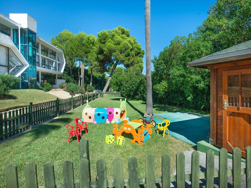 Bunter Spielplatz mit großen Würfeln, tierförmigen Figuren und Holzhaus vor grüner Naturkulisse.