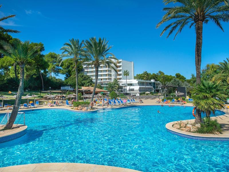 Großer blauer Pool surrounded by Palmen und Liegestühlen, Hotelgebäude im Hintergrund bei klarem Himmel.