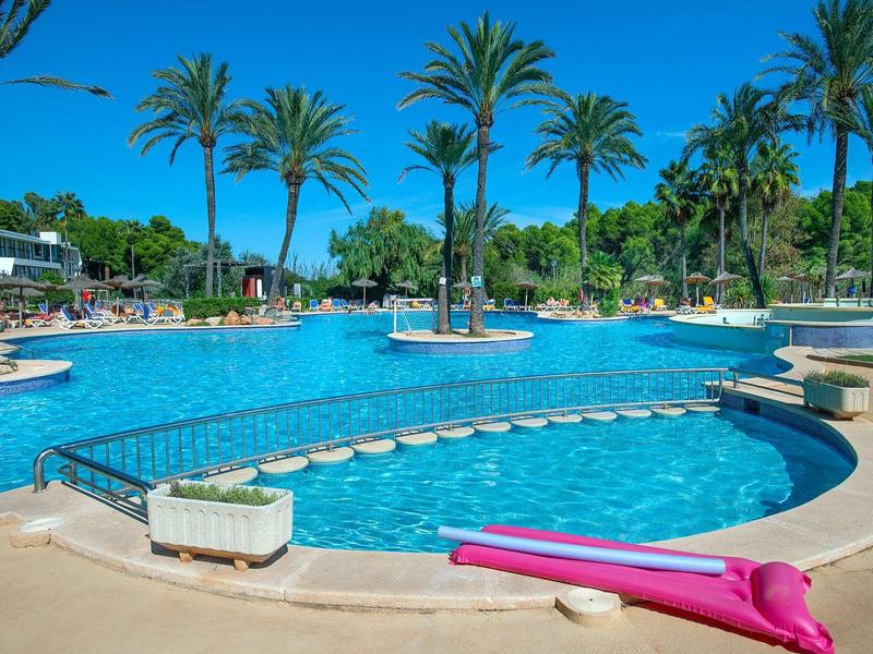 Großer, blauer Pool mit Palmen, Inseln und pinker Luftmatratze bei strahlend blauem Himmel.