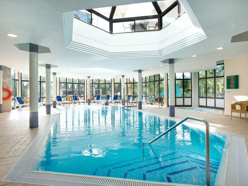 Hell beleuchtetes Hallenbad mit großem Pool, vielen Stühlen und großen Panoramafenstern.